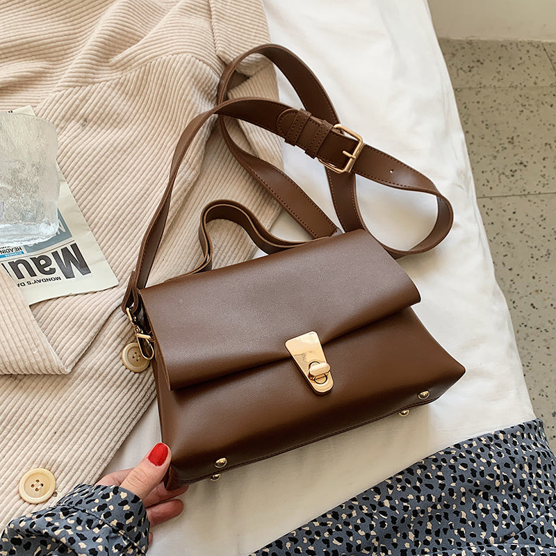 Brown leather messenger bag with gold accents held by a hand on light-colored surface.