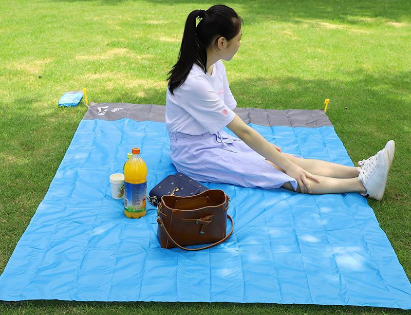 Woman sitting on a blue picnic blanket with a bottle and bag in a grassy area.