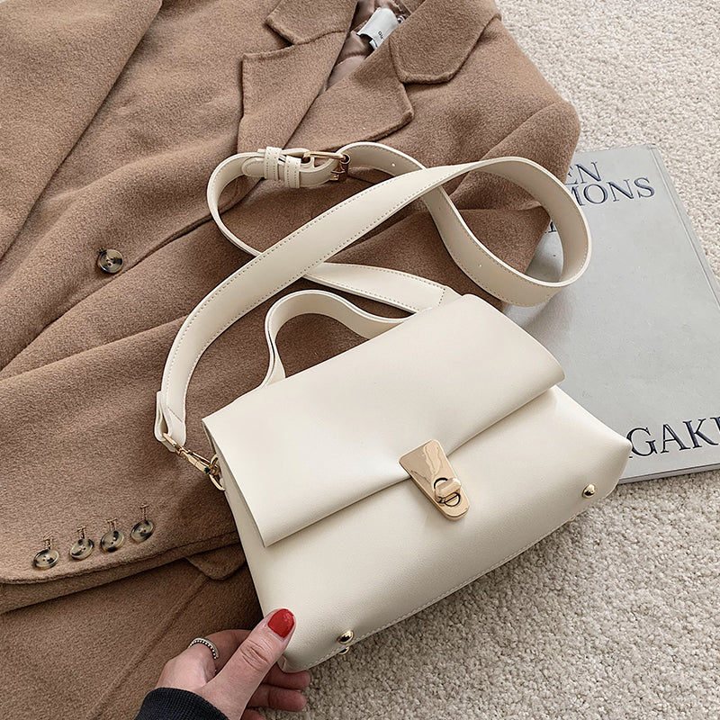 White leather messenger bag held by a hand with red nail polish, placed on a beige surface with a brown coat and magazine in the background.