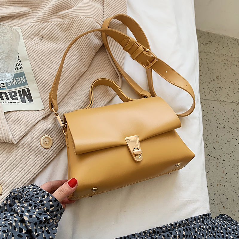 Yellow leather messenger bag held by a hand with red nail polish on a textured surface.